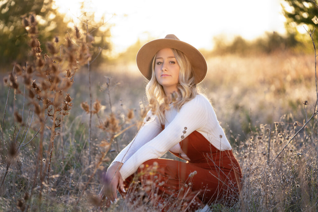Senior girl in a tan wide-brim hat, cream sweater, and rust overalls sitting in a golden field at sunset for fall senior pictures at Northwest Community Park in Fort Worth