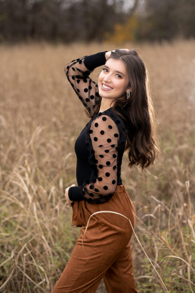 Senior girl in a black polka dot top and rust pants standing in a tall brown grass field for fall senior pictures in Keller, Texas field
