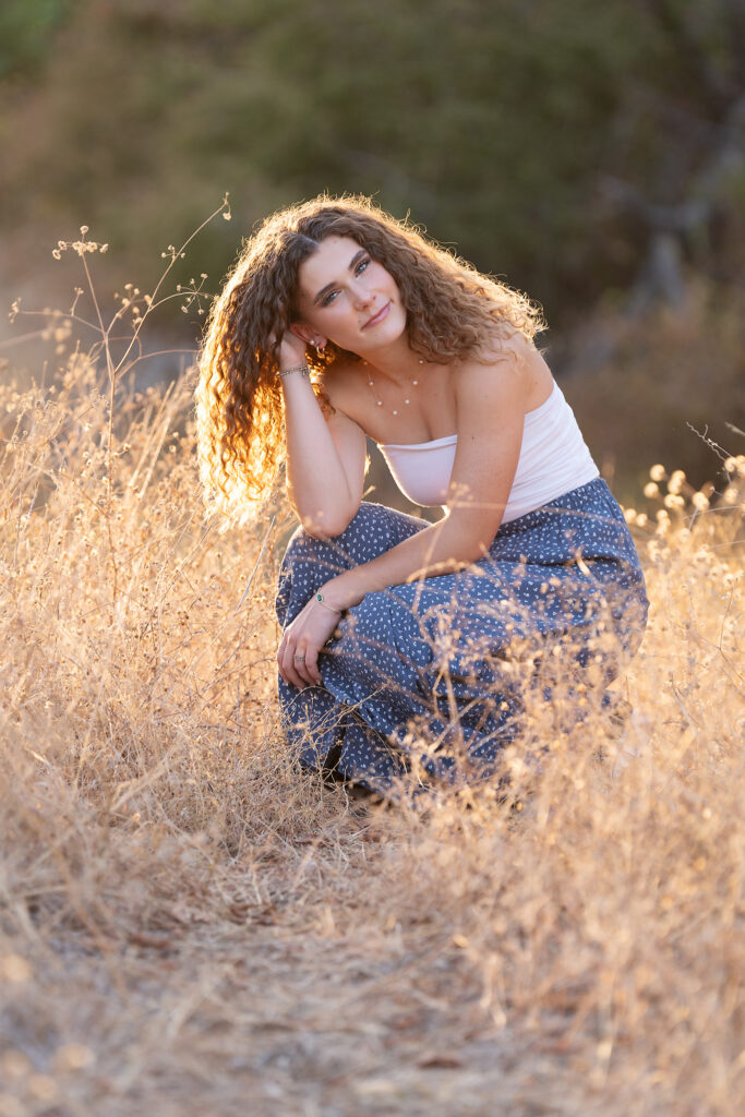 Senior girl with curly hair crouching in a warm, glowing golden field at sunset for fall senior pictures in Dallas-Fort Worth Northwest Community Park