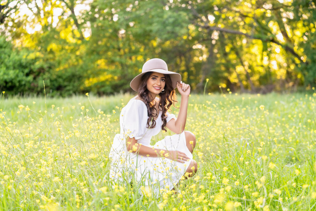 Senior girl in a white dress and tan wide-brim hat sitting in a field of yellow wildflowers for spring senior pictures in Dallas-Fort Worth