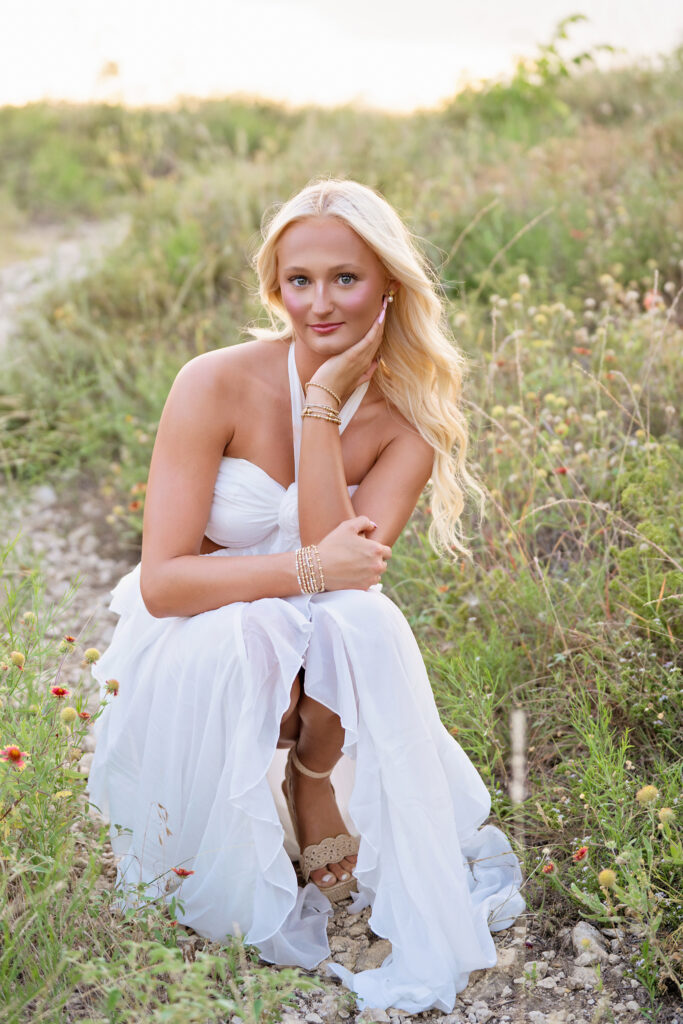 Senior girl in a white dress sitting among summer wildflowers during an evening senior session at Tandy Hills Nature Center in Fort Worth